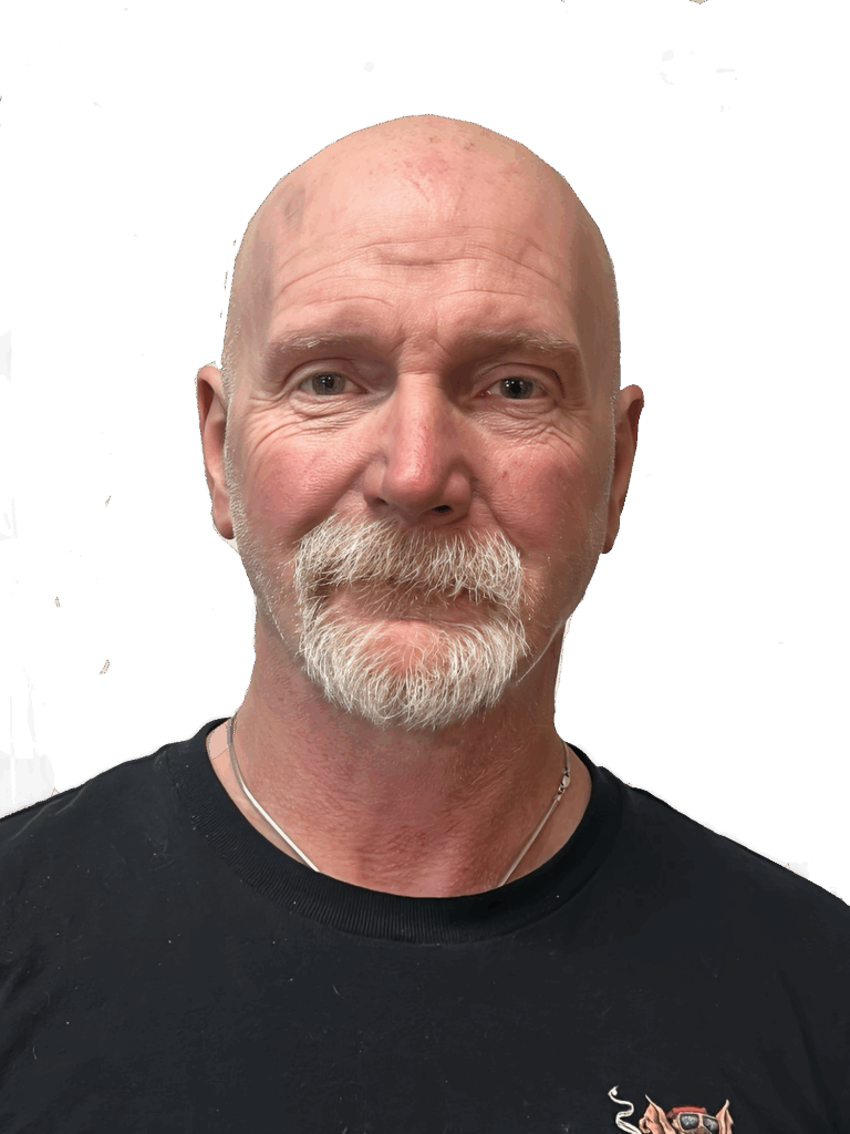 A bald man with a mustache and beard is wearing a black shirt and a silver necklace, set against a plain background.