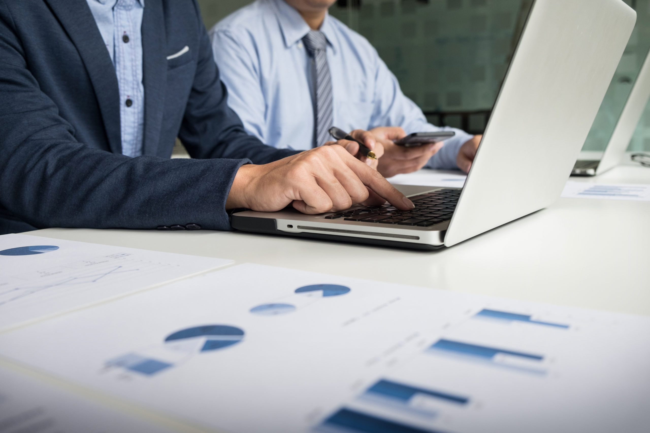 Two individuals in formal attire work on a laptop at a desk with business charts.