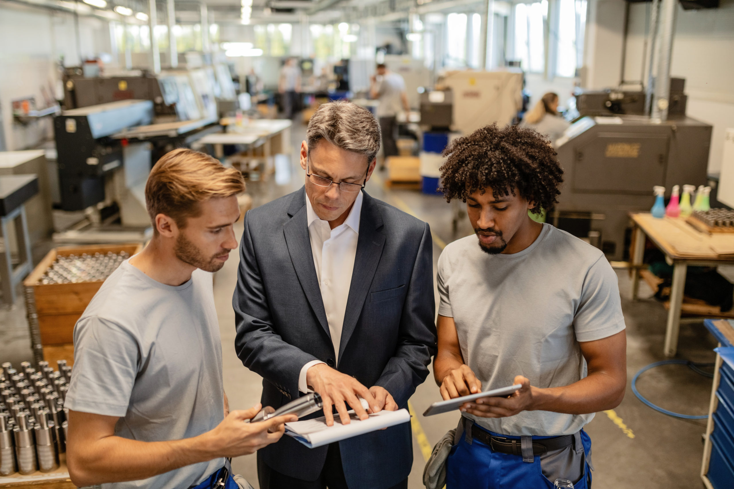 A group of people is engaged in discussion in an industrial workspace, examining documents and tablets.
