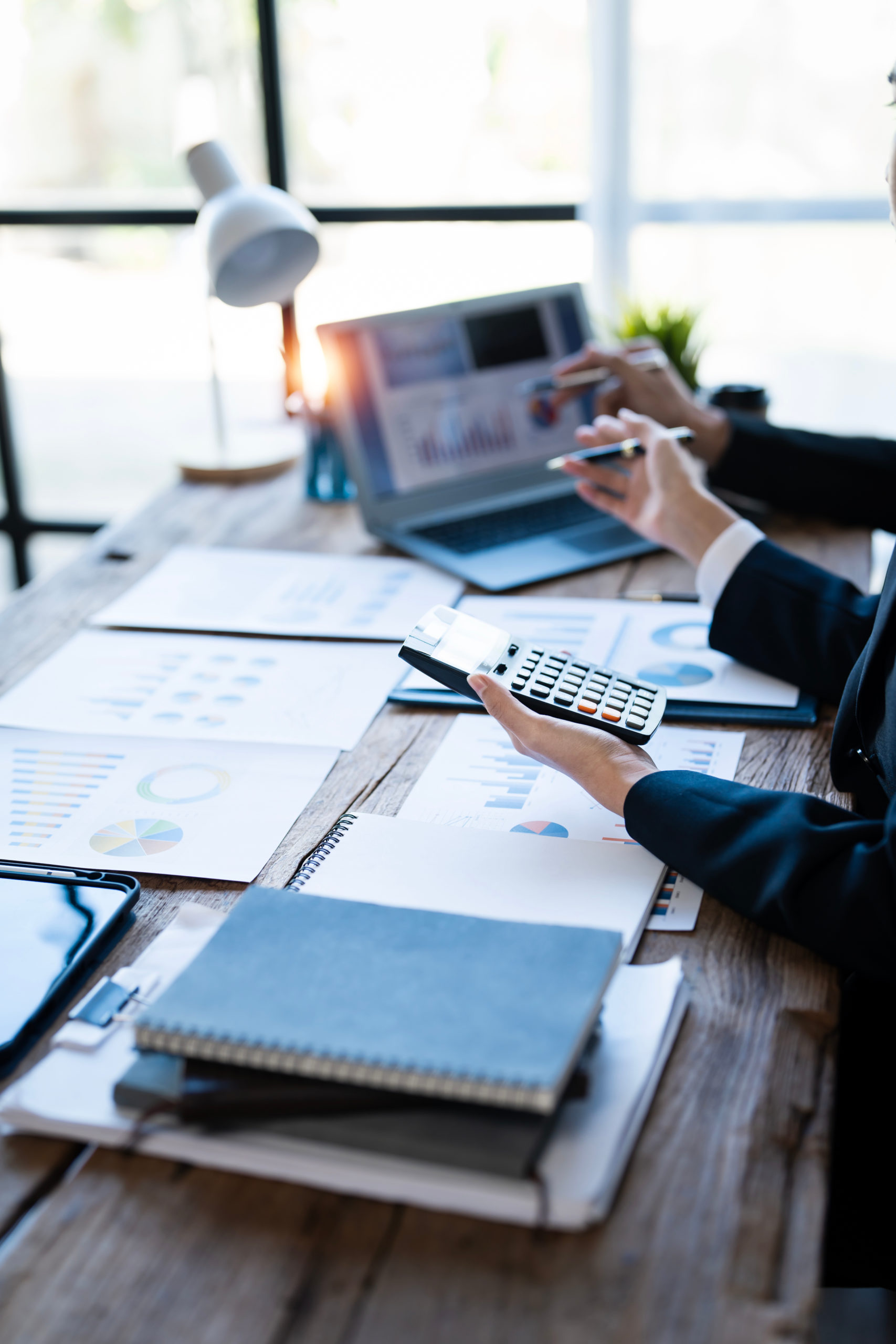 Individuals are engaged in reviewing financial documents and using a laptop and calculator at a wooden desk.