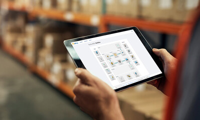 A person is holding a tablet displaying a warehouse map or inventory list in a storage area with shelves and boxes.