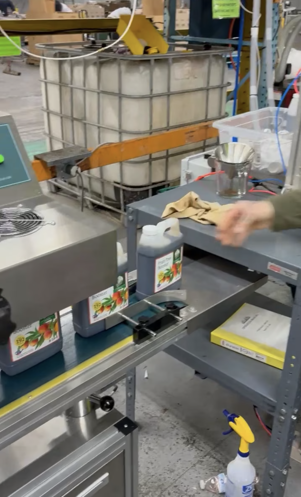 A hand in motion near a conveyor belt with containers and a display screen in a factory setting.